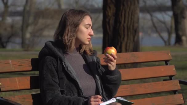 小女孩坐在公园的长凳上, 吃苹果, 在笔记本上写字。 — 图库视频影像
