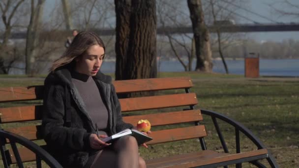 Chica bonita come manzana en el banco en el parque, y hace notas en el cuaderno — Vídeo de stock