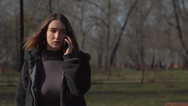 Молода дівчина розмовляє по телефону, ходячи в парку — стокове відео