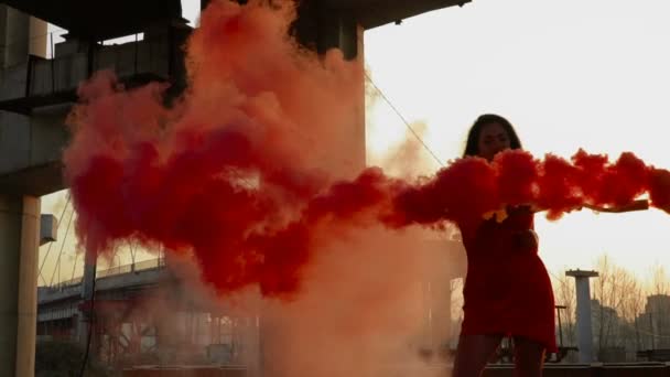 Junge Frau in rotem Kleid tanzt mit rotem Rauch nahe verlassener Brücke — Stockvideo