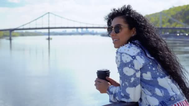 Hermosa morena en gafas de sol disfruta de la vista sobre el río y bebe café — Vídeos de Stock