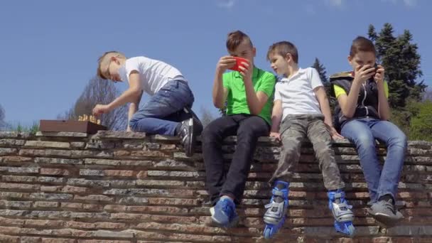 Четыре подростка сидят у бетонной стены в парке — стоковое видео