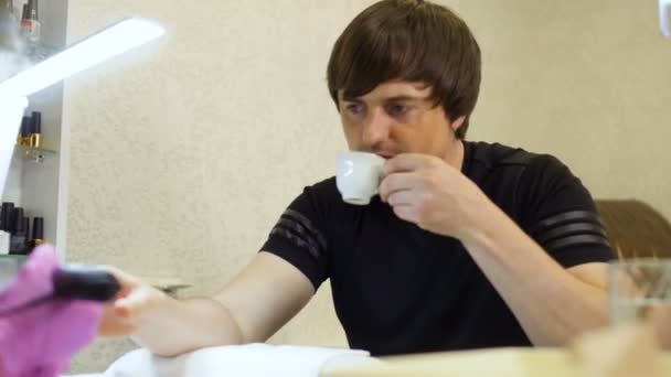 Молодой человек расслабляется и пьет кофе во время процедуры маникюра — стоковое видео