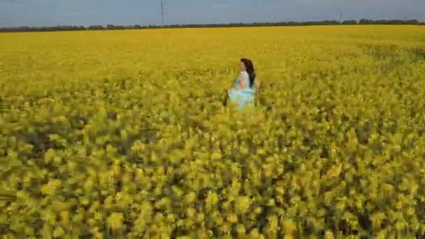 Vacker kvinna i blå klänning kör igenom ett gult fält med raps blommor — Stockvideo