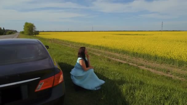 若い女性が彼女の車の近くに道端に座るし、美しい菜種の花畑をお楽しみください。 — ストック動画