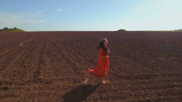 Jonge zorgeloos vrouw in lange rode jurk draait op geploegd veld — Stockvideo