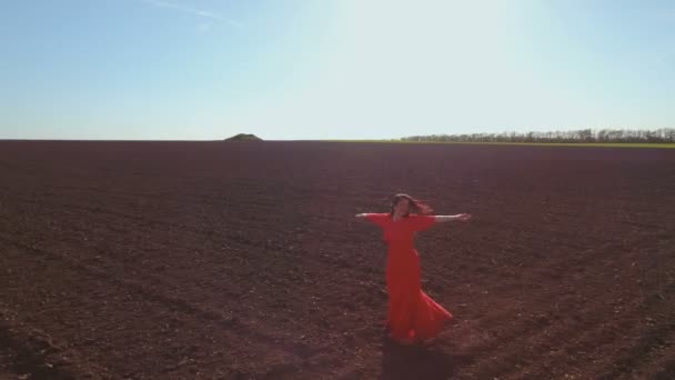 Glückliche unbeschwerte Frau im langen roten Kleid, die sich auf gepflügtem Feld umdreht — Stockvideo