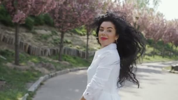 Очаровательная улыбающаяся женщина гуляет в парке среди аллеи с цветущими деревьями — стоковое видео