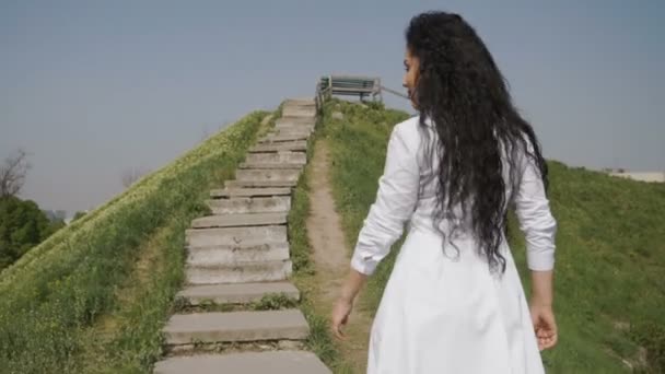 Elegante Frau im langen weißen Kleid bewegt sich die Treppe auf dem Hügel hinauf — Stockvideo