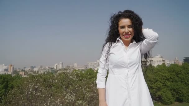 Красивая женщина в белом платье позирует перед камерой на фоне зеленого города — стоковое видео