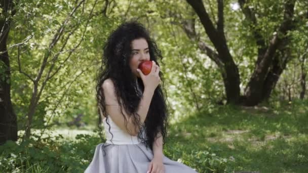 Piękna Brunetka siedzieć w ogrodzie letnim i jeść Czerwone jabłko — Wideo stockowe