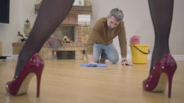Erişkin beyaz adamın portresi zemini yıkıyor ve karısıyla ya da kız arkadaşıyla konuşuyor. Ön planda zarif kırmızı yüksek topuklu ayakkabılar giyen güzel kadın bacakları. Suçlu kılıbık ev işi yapıyor..
