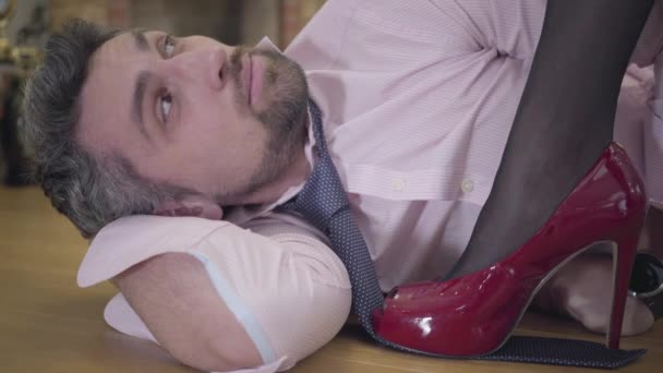 Detailní záběr pohledného šedovlasého bělocha ležícího na podlaze. Krásná samičí noha v červených botách na vysokém podpatku, stojící na kravatě. Portrét usměvavého poskoka. — Stock video