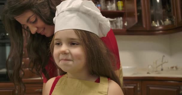 Μια ενήλικη λευκή γυναίκα βάζει ποδιά σε ένα χαριτωμένο κορίτσι με λευκό καπέλο και τη φιλάει. Κοντινό πλάνο πορτρέτο μιας ευτυχισμένης κόρης με μακριά καστανά μαλλιά που βοηθάει τη μαμά με το μαγείρεμα. Κινηματογράφος 4k πλάνα Prores Hq. — Αρχείο Βίντεο