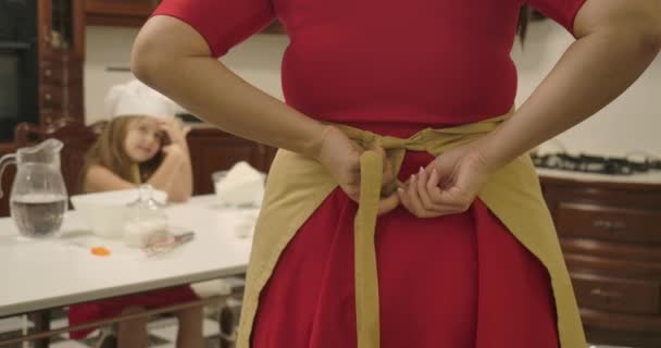 Back view of Caucasian woman in red dress tying apron. Blurred little girl sitting at the table at background and looking at lovely mother. Happy family cooking together. Cinema 4k footage ProRes HQ. — Stock Video
