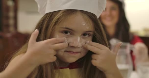 小麦粉と顔に線を引く喜びの白人の少女のクローズアップ肖像画。彼女の若い笑顔の母親は背景に立っている。幸せな家庭料理を一緒に。映画4k映像｜Prores Hq. — ストック動画