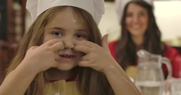 Zbliżenie twarzy wesołej białej dziewczynki rysującej linie na twarzy mąką i mruganiem. Młoda uśmiechnięta matka stojąca w tle. Szczęśliwa rodzinna kuchnia. Kino 4k materiał Prores Hq. — Wideo stockowe