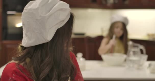 Młoda kaukaska matka spogląda wstecz na córkę mieszającą składniki do piekarni, zwracając się do kamery i uśmiechając się. Portret dziewczyny w kapeluszu kucharza w kuchni. Kino 4k materiał Prores Hq. — Wideo stockowe