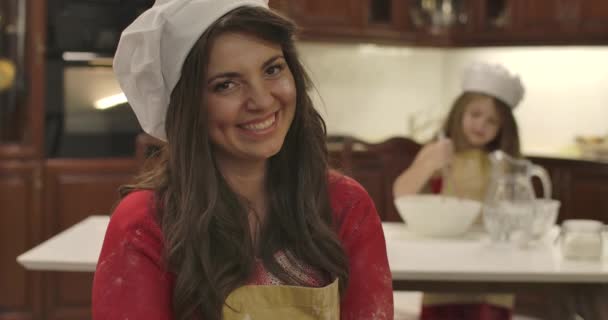 Joven mujer caucásica sonriente mirando a la cámara y volviendo a su pequeña hija mezclando ingredientes para la panadería. Retrato de mujer en sombrero de cocinero en la cocina. Cinema 4k metraje ProRes HQ . — Vídeos de Stock