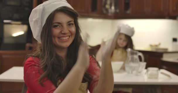 Primer plano de la joven mujer caucásica mirando a la cámara, aplaudiendo, harina volando alrededor. Retrato de mujer sonriente en sombrero de cocinero, hija mezclando ingredientes en el fondo. Cinema 4k metraje ProRes HQ . — Vídeo de stock