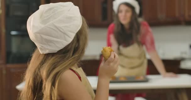 Ung kaukasisk mor tittar på hennes underbara barn äta bageri i förgrunden och skicka luft kyssar. En liten vit flicka som tuggar god mat. En söt liten flicka som äter bröd hemma i köket. — Stockvideo