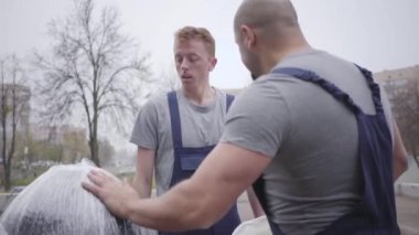 Sakallı beyaz adam genç işçiye beyaz koruyucu kask takıyor, yeni çalışan paketi alıyor ve uzaklaşıyor. Taşıyıcılar işte..