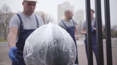 Beyaz kasklı ve üniformalı yetişkin bir adam paketi alıyor ve binaya doğru yürüyor. Kaslı teslimatçı iş başında. Kamera aşağı iniyor.