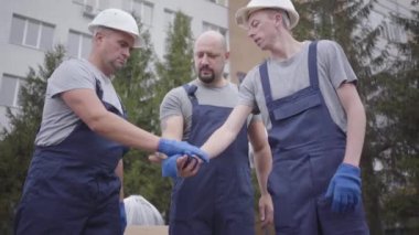 Üç genç Kafkasyalı işçi el ele tutuşuyor, başlarını sallıyor ve çalışmaya başlıyorlar. Beyaz miğferli iki nakliyeci paketleri alıp binaya gidiyor..