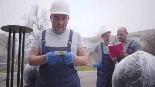 Hombre caucásico feliz con casco blanco y ropa de trabajo azul contando dinero y sonriendo. Al repartidor le pagaron por su trabajo. Sus colegas revisan los documentos en segundo plano . — Vídeos de Stock