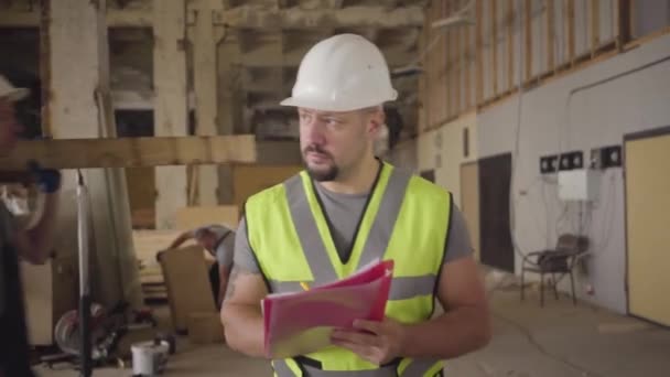 Retrato de cerca del trabajador caucásico barbudo en casco blanco caminando en el sitio de construcción, mirando a su alrededor y escribiendo en documentos. Otros constructores trabajando en segundo plano. Supervisor de trabajo de verificación . — Vídeo de stock