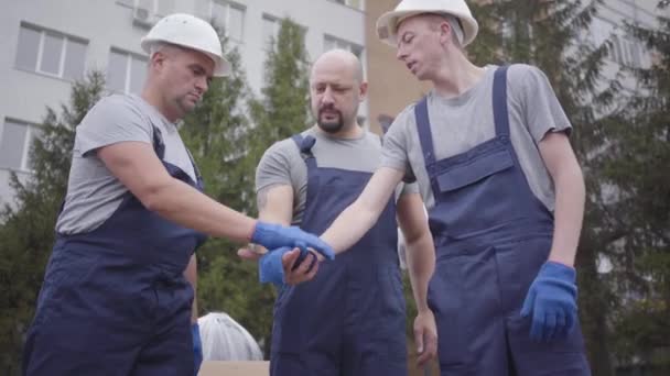 Tres jóvenes trabajadores caucásicos poniéndose las manos encima, agitando cabezas y comenzando a trabajar. Dos movedores en cascos blancos tomando paquetes e yendo al edificio, supervisor llenando documentos . — Vídeo de stock