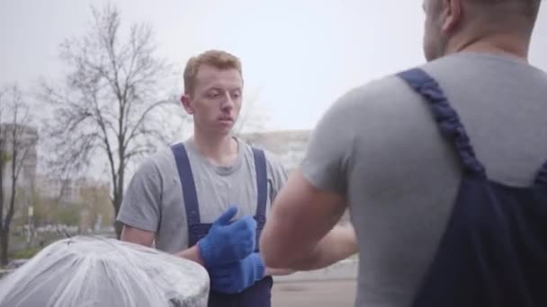 Muscular Caucasian supervisor putting white protective helmet on young worker, new redhead employee taking package and walking away. Chief talking and gesturing at the background. Movers at work. — Stock Video