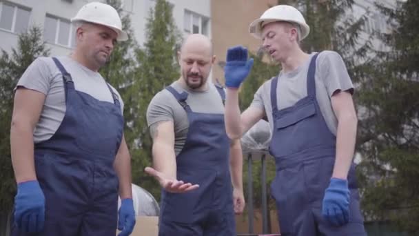 Três movimentadores musculosos caucasianos a porem as mãos uns nos outros e a começarem a trabalhar. Trabalhadores de uniforme azul levando pacotes e indo para o prédio, supervisor de preenchimento de documentos. Câmara a mover-se . — Vídeo de Stock