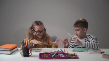 Beyaz bir erkek ve kız masada oturuyor ve renkli kalemlerle kavga ediyorlar. Evde oynayan neşeli çocuklar var. Erkek ve kız kardeş okuldan sonra eğleniyor..