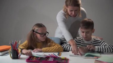 Nazik, beyaz bir anne çocuklarına ödevlerinde yardım ediyor. Gözlüklü küçük tatlı kız ve erkek kardeşi masada oturup egzersiz kitaplarına yazıyor. Kardeşler evde ders çalışıyor..