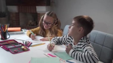 Egzersiz kitaplarına yazı yazan ve konuşan iki beyaz çocuğun portresi. Gözlüklü bir kız ve okulda masada oturan bir oğlan. Kamera soldan sağa hareket ediyor.