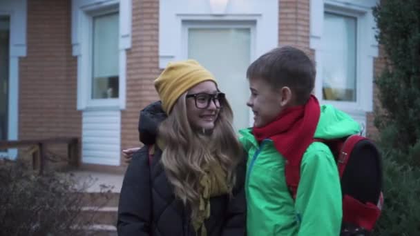 Liten vit pojke i grön rock kramar söt flicka i glasögon och gul hatt bredvid det stora huset. Positiv bror och syster med ryggsäckar som lämnar skolan. — Stockvideo