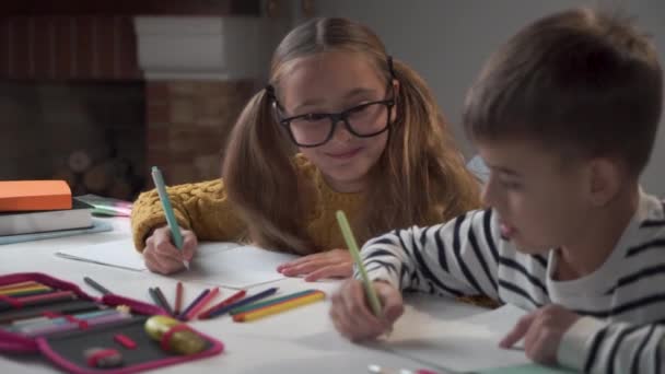 Dos compañeros de escuela caucásicos sentados a la mesa y escribiendo en libros de ejercicios. Chica con gafas de vista mirando al chico y sonriendo. Compañeros de clase haciendo tareas en la escuela . — Vídeo de stock