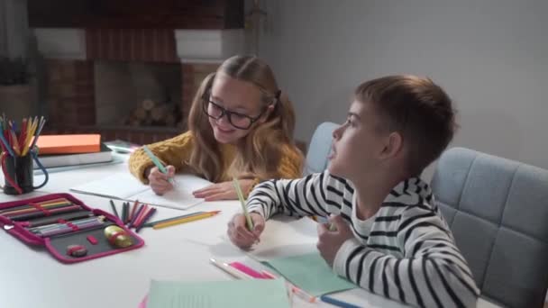 Retrato de dos alegres niños caucásicos escribiendo en libros de ejercicios y hablando. Chica en gafas y niño sentado en la mesa en la escuela y haciendo tareas. Cámara moviéndose de izquierda a derecha . — Vídeo de stock