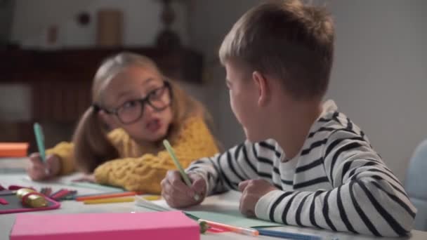 Detailní portrét usměvavého bělocha v pruhovaném saku, jak píše do sešitu. Rozmazaná dívka v brýlích mluví v pozadí. Školáci studující doma nebo ve škole. — Stock video