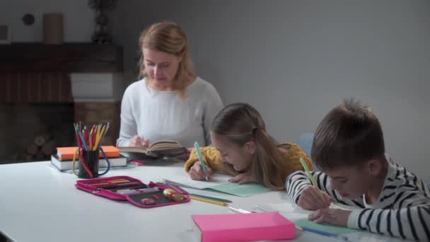 Jonge blanke vrouw die boek leest en kinderen controleert om in oefenboeken te schrijven. Jongen en meisje doen huiswerk met moeder. Broers en zussen die thuis studeren. Onderwijs, intelligentie, basisschool. — Stockvideo