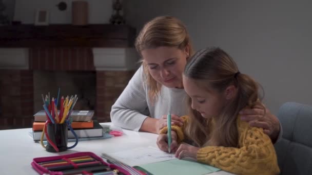 幸せな白人の母親は、彼女のかわいい娘が宿題をしているのを見て、笑顔で抱きかかえています。小賢しい少女の練習帳に書いています。子供の勉強を手伝ってくれる幸せな女性。教育. — ストック動画