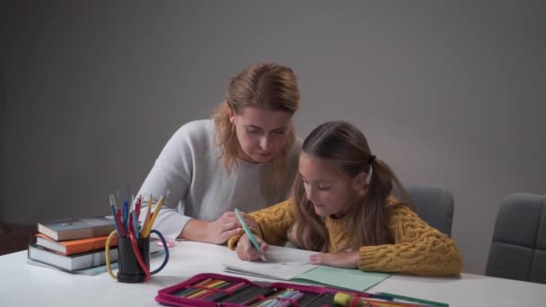 Маленька кавказька дівчинка робить домашнє завдання з мамою. Молода жінка пояснює дочці, що зробила помилки. Батьки допомагають школярці вчитися. Початкова школа, домашня освіта. — стокове відео
