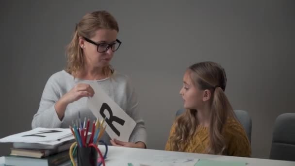 Portret pięknej białej dziewczyny uczącej się alfabetu z matką. Młoda inteligentna kobieta w okularach pokazująca córce kartkę z literą R i wyjaśniająca prawidłową wymowę. — Wideo stockowe