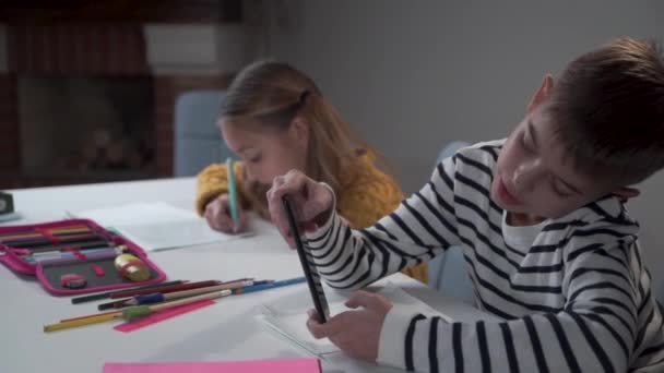 Två vita barn som sitter vid bordet i skolan. flitig flicka med två hästsvansar skriver i träningsboken som stygg pojke spelar spel på smart telefon. Kameran rör sig från vänster till höger. — Stockvideo