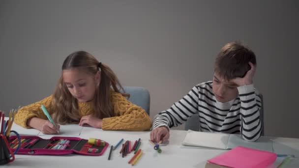 Sad Caucasian boy and happy girl sitting at the table at school or at home. Diligent smart sister doing homework as her bored brother playing with pens. Different attitude to the education, behaviour. — Stock Video