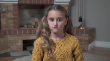 Hardallı süveter giyen, göğsünde el ele tutuşan ve başını sallayan üzgün, beyaz bir kızın yakın çekimi. Evdeki şöminenin önünde oturan üzgün çocuk..