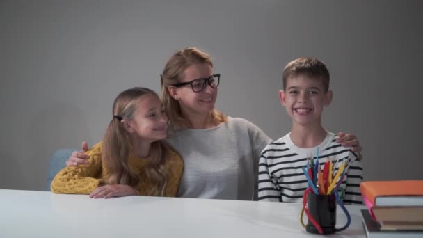 Dos pequeños niños caucásicos sonrientes posando con su inteligente maestra sentada a la mesa. Hermosa tutora en anteojos abrazando a niña y niño y sonriendo. Concepto educativo . — Vídeos de Stock