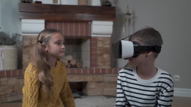 귀여운 백인 자매가 오빠가 VR 안경을 쓰고 놀기를 기다리고 있습니다. 형제들은 집에 있는 벽난로 앞에 앉아 있습니다. 주말, 여가 활동, 휴식, 어린 시절. — 비디오