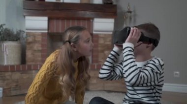 VR gözlüklü yakışıklı beyaz çocuk güzel kızla konuşuyor ve etrafa bakınıyor. Erkek ve kız kardeş evde yeni bir aletle oynuyorlar. Hafta sonları, boş zaman aktiviteleri, dinlenme, çocukluk..